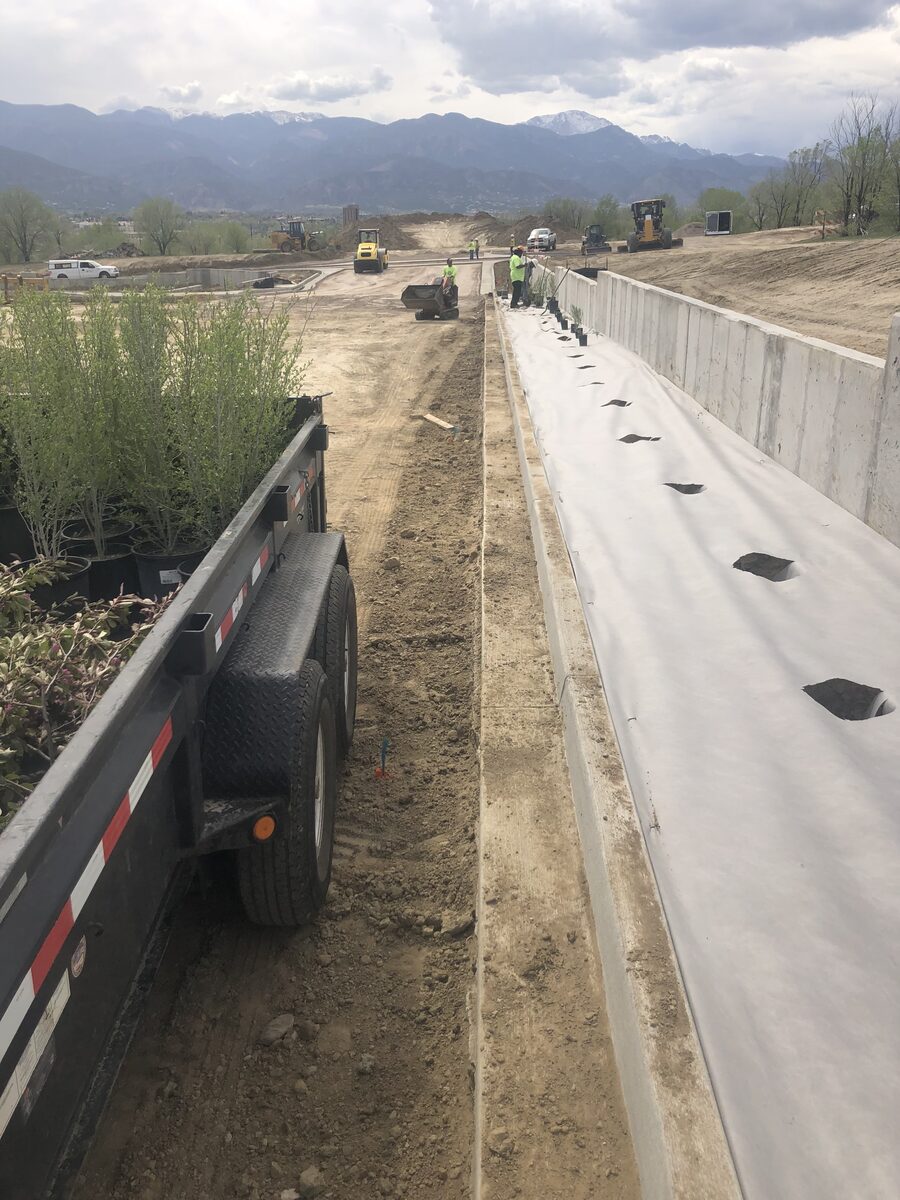 Plant material staged on a trailer beside a new curb and retaining wall while graders work the corridor behind