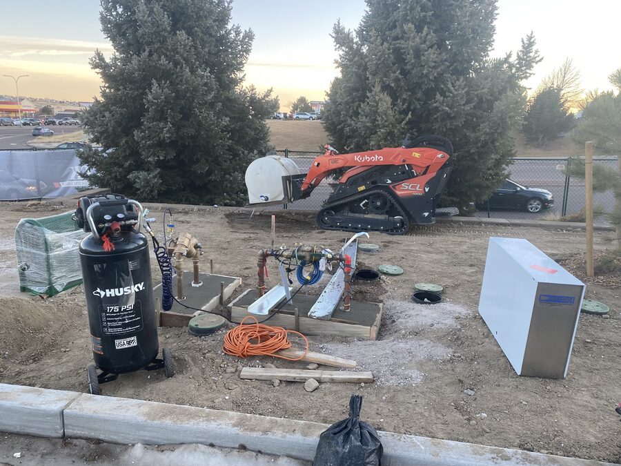 Irrigation manifolds set in concrete beside a compact track loader and air compressor at a commercial site